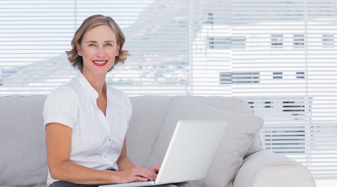 Eine blonde Dame sitzt auf einem grauen Sofa und hält ein Laptop auf den Knien fest.